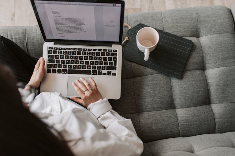 From above of woman in casual wear sitting on comfortable couch with cup and typing on keyboard of laptop while working remotely in cozy living room